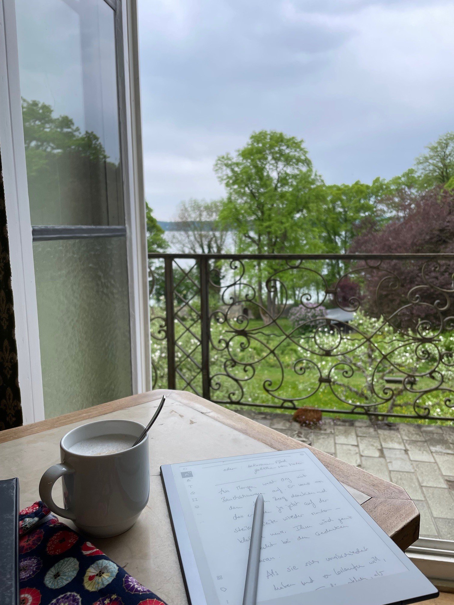 Blick aus dem Fenster auf einen Garten, dahinter der Starnberger See. Im Vordergrund eine Tasse und ein Schreibtablet mit Stift