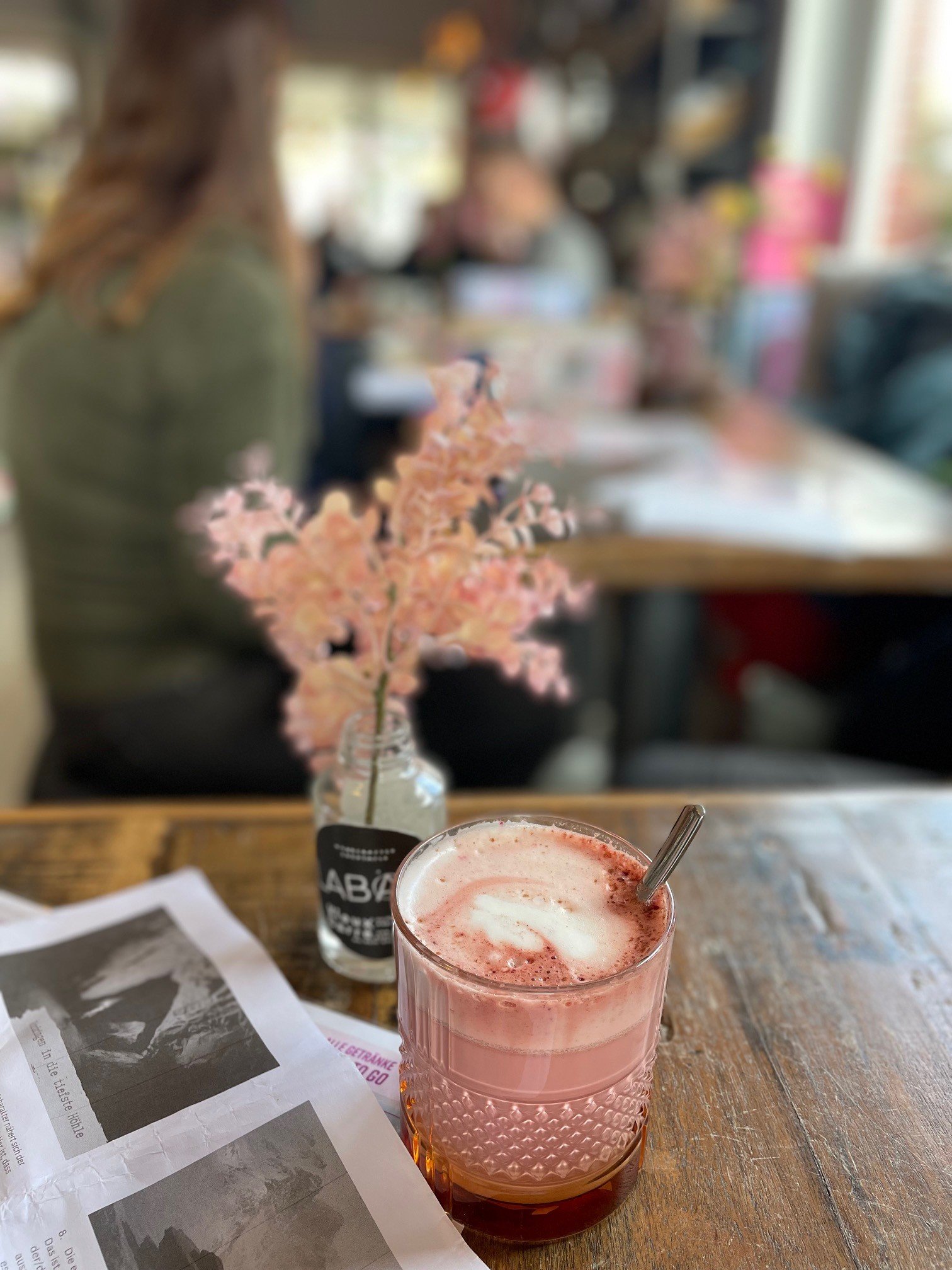 In einem Café: Rosa Getränk in Tasse und Rosa Trockenblume in Vase auf Tisch. Daneben ein paar Blätter zur Heldenreise. Im Hintergrund Menschen, die schreiben und Kaffee trinken.